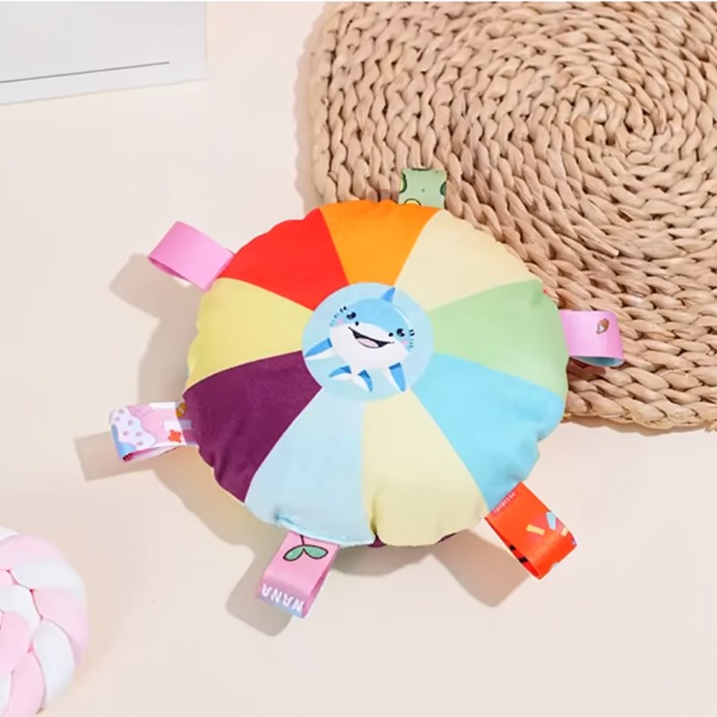 Bunte, runde Plüschhundeball mit Klingel liegt auf einer beige gewebten Matte.